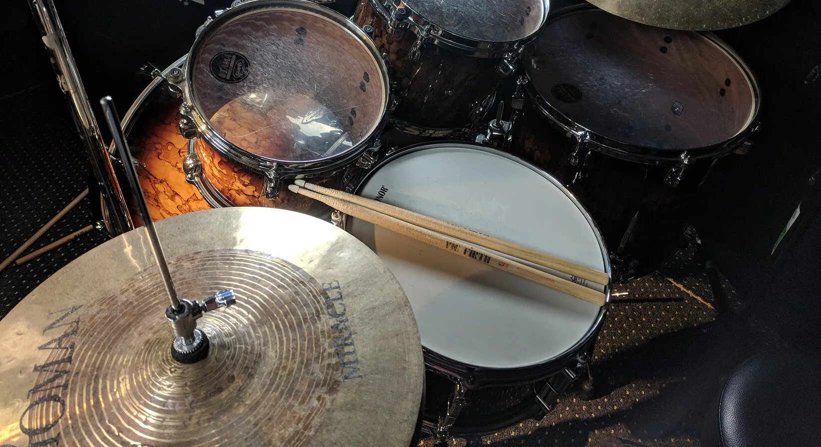 Drumsticks resting on a snare drum next to a hi-hat cymbal, seen from the drummer's seat