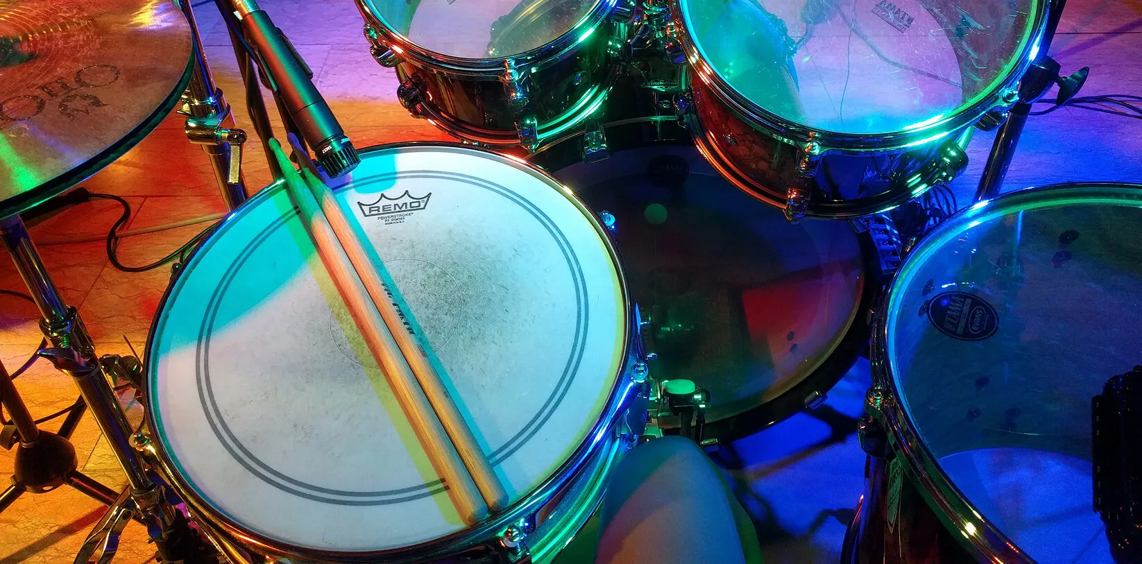 Drumsticks resting on a snare drum under colourful stage lights, with toms and a microphone around it